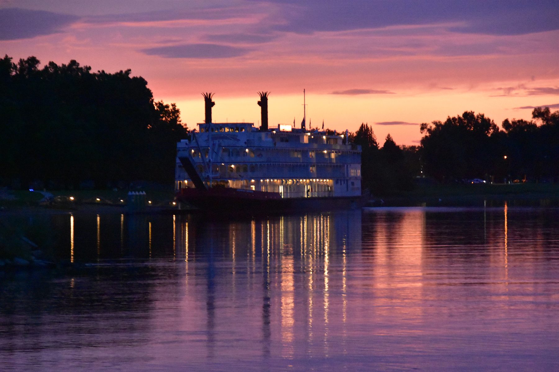 Paddlewheel riverboat with its lights on, moored at dusk under a pink sky
